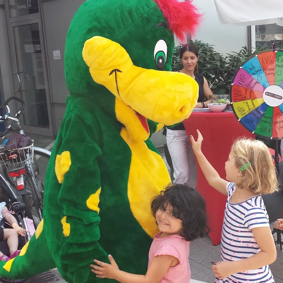 Kinder begrüßen Drache Dinoli an einem Glücksrad-Stand auf einem Fest