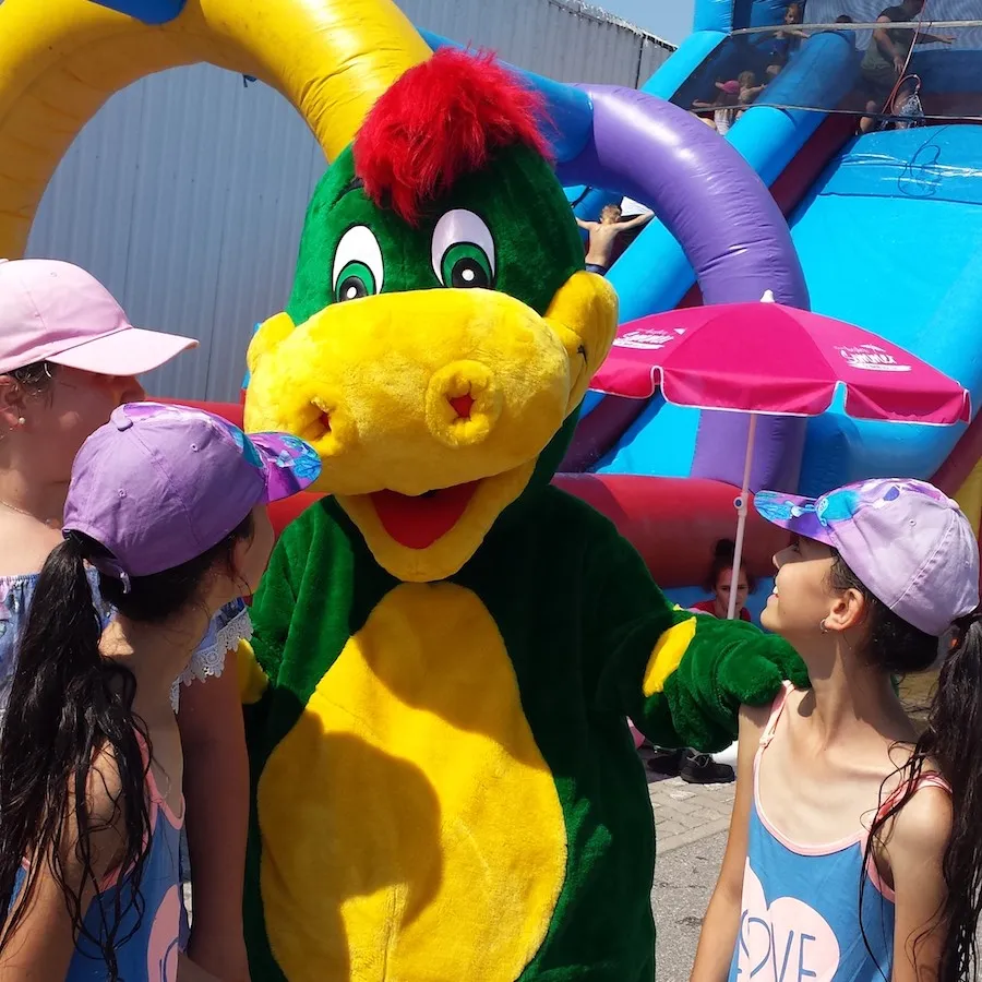 Drache Dinoli umringt von Kindern mit Sonnenkappen auf einem Sommerfest