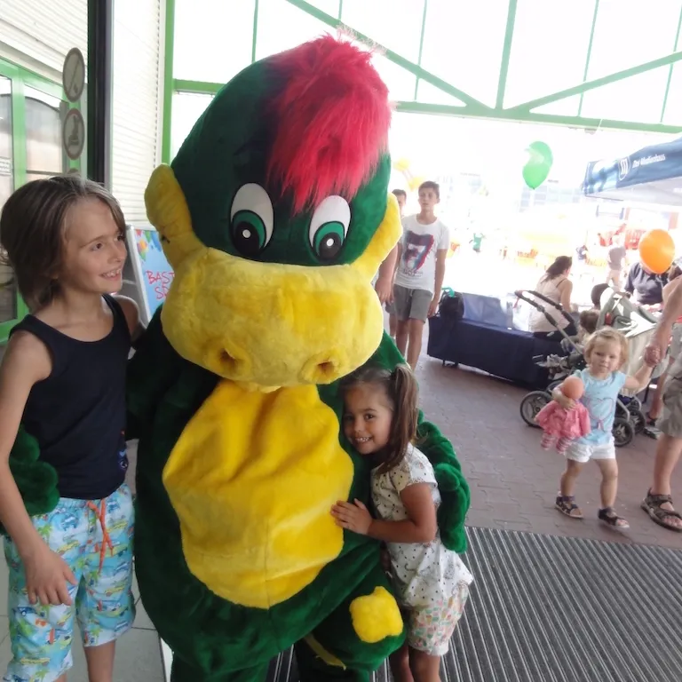 Drache Dinoli wird auf einem Fest von zwei lachenden Kindern umarmt