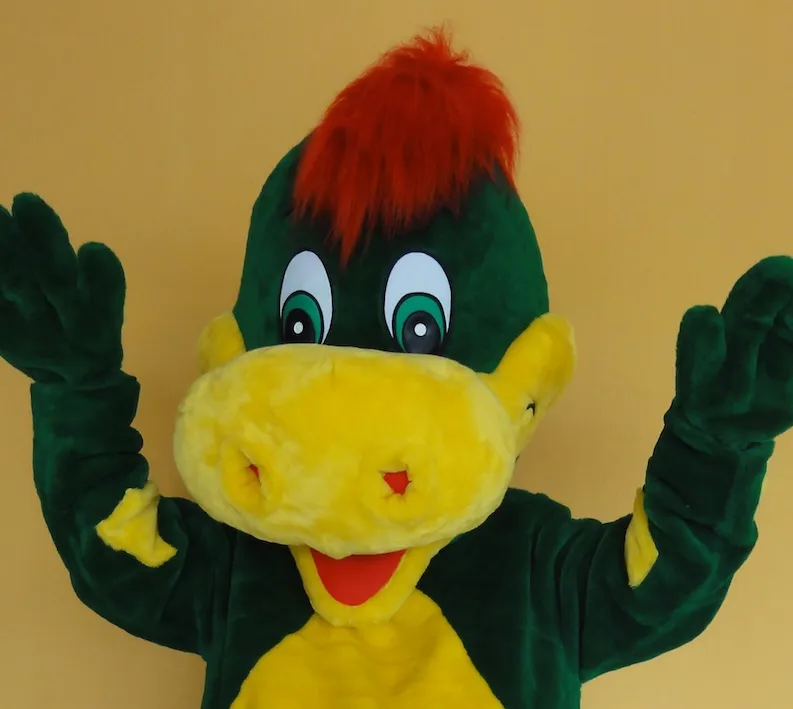 Maskottchen Drache Dinoli reißt vor einer gelben Wand fröhlich die Arme hoch